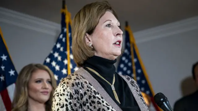 黒いハイネックのトップスにヒョウ柄のカーディガンを着たシドニー・パウエル氏が、前方を見て話をしている。後ろにはジェナ・エリス氏が微笑んで立っている