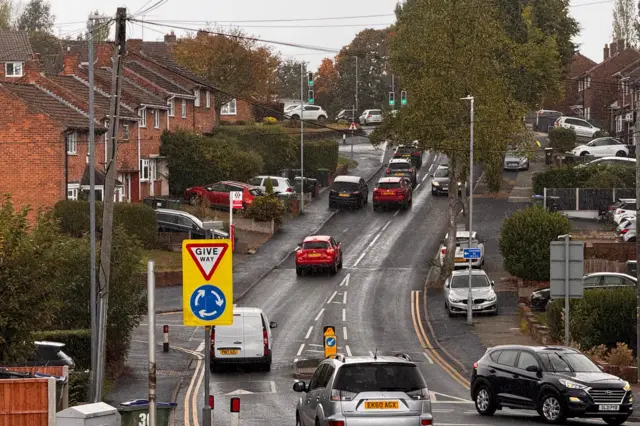 Cars driving up and down a road in a residential area. A bus stop and a 'give way' sign are on the left-hand side of the road.