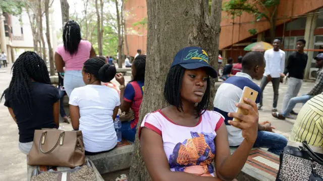 Aworan awọn akẹkọọ to jokoo sita lai ṣiṣẹ ninu ọgba ile ẹkọ giga. Akẹkọọbinrin to wa niwaju ninu aworaN naa n yẹ foonu rẹ wo