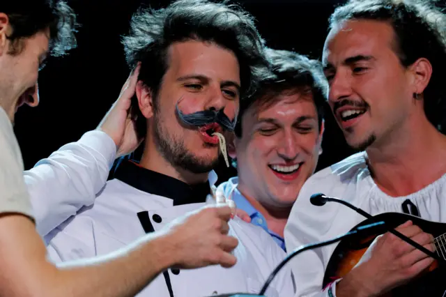 Четверо мужчин в белом стоят рядом друг с другом, один из них засаывает макароны, еще один играет на гитаре.
