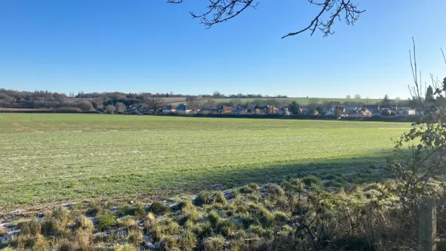 Una imagen de un campo verde en Redbourn. Hay un poco de escarcha en el suelo al frente de la imagen y luego un campo verde con una hilera de casas detrás. El cielo es azul detrás de las casas.