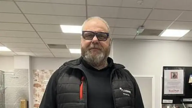 Michael King está de pie en una habitación, sonriendo a la cámara. Tiene el pelo corto y canoso, lleva gafas y un abrigo negro.