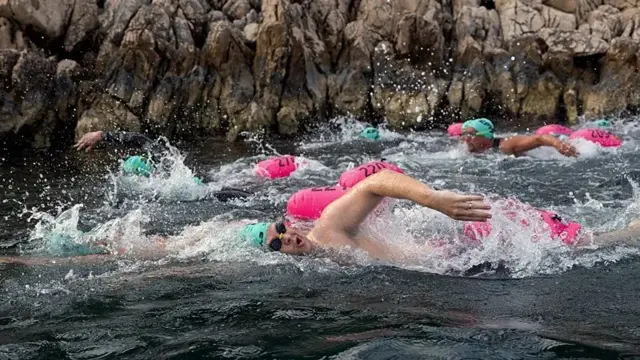 Pessoas com coletes e óculos de natação nadando. Há boias rosas ao lado.