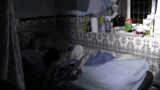 One of the rooms used for sexual exploitation and raided by the team, showing an unmade bed with a blue cover and a shelf next to a window full of jars and objects.