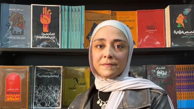 سيدة ترتدي حجاباً أبيض وتنظر إلى الكاميرا وهي تقف أمام أرفف معروض عليها العديد من الكتب