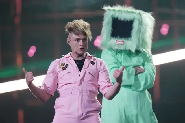 Look Mum No Computer in a pink boiler suit with a dancer in a green one and a fluffy box on their head behind him