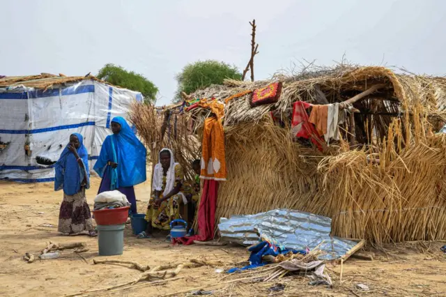 Certaines femmes sont debout, d'autres assises, avec à proximité des objets de première nécessité tels que des seaux et des casseroles. Leurs expressions et leur langage corporel suggèrent un mélange de lassitude et de résilience.