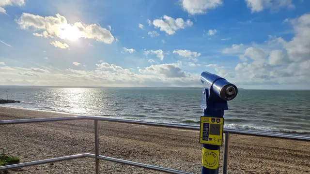 Un telescopio junto a la playa ofrece vistas a las arenas amarillas y a un mar de aspecto tranquilo con el sol reflejándose en el agua.