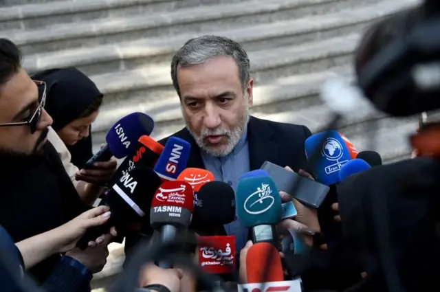 Arakçi resmi giyimi merdivenlerin bitiminde ona yakın mikrofona konuşuyor, solunda güneş gözlüklü erkek arkasında türbanlı bir kadın var. 