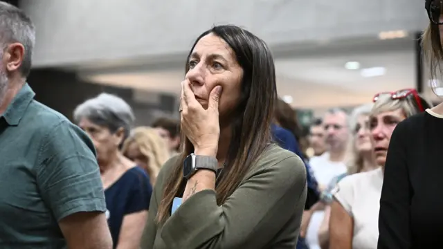 Žena duge smeđe kose, drži ruku na ustima, ima crni sat, u zelenom džemperu