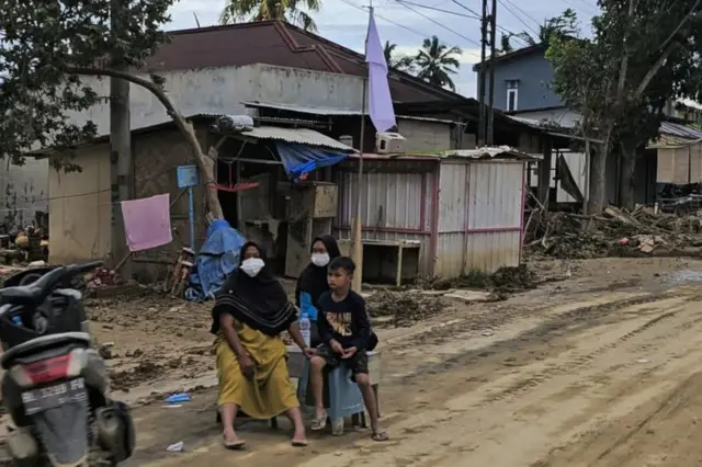 Warga dan anak-anak duduk di bawah bendera putih yang terpasang di sisi Jalan Lintas Sumatera, Aceh Tamiang, Senin (15/12)