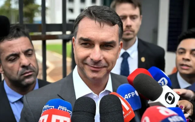 Foto mostra Flávio Bolsonaro cercado por microfones usando um terno escuro