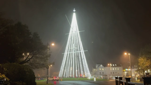 La foto muestra un árbol de Navidad blanco iluminado en St. Peter Port en la oscuridad. Hay farolas encendidas y se ven coches en la rotonda.