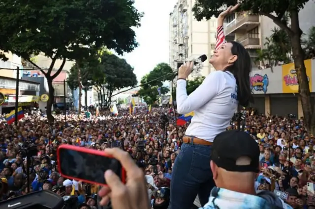 Corina discursando no microfone e sorrindo ao olhar pra cima; ela é ouvida por centenaspré aposta esportivapessoas na rua