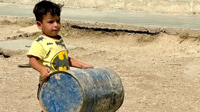 A young boy in a Batman t-shirt carrying an empty metal barrel outside