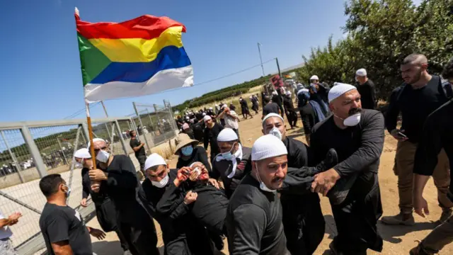 Um membro da comunidade drusa segura sua bandeira enquanto carrega um manifestante ferido durante um protesto na vila de Majdal Shams, nas Colinas de Golã anexadas por Israel, em 21 de junho de 2023, contra um projeto de turbinas eólicas israelenses supostamente planejado para as terras agrícolas da vila.