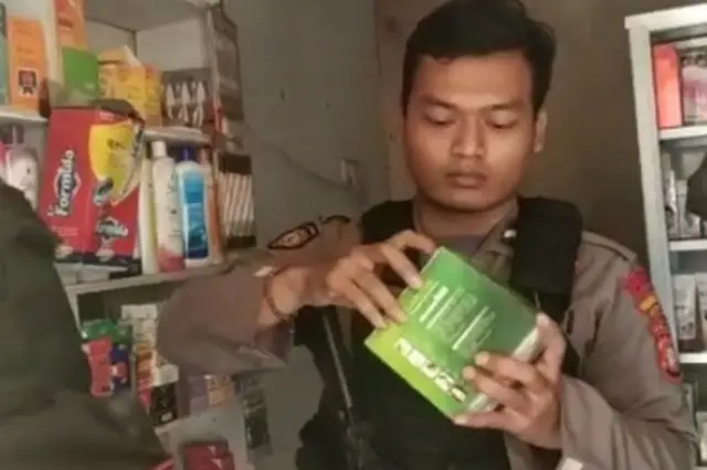 Seorang polisi sedang memegang kotak berisi obat keras dengan latar belakang toko kosmetik.