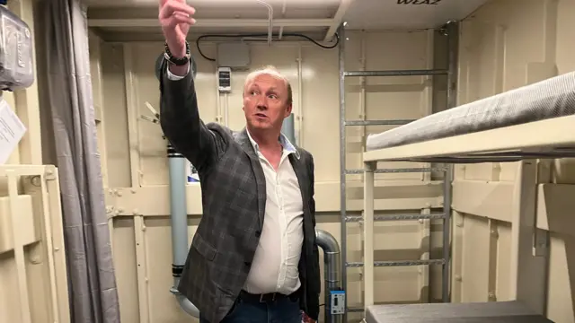 Janusz, de jeans e jaqueta xadrez, aponta para o teto de um de seus abrigos. O local tem um beliche e um vaso sanitário com cortina. É limpo, mas espartano.

