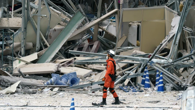Un bombero, con uniforme anaranjado, botas negras, casco y mascarilla blancos, pasa por el lado de los escombros de una infraestructura

