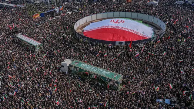 Pessoas acenam com bandeiras nacionais iranianas enquanto participam dos funerais de comandantes da Guarda Revolucionária Islâmica (IRGC), do exército e de outros mortos nos primeiros dias dos ataques dos EUA e de Israel contra o Irã, na Praça Enghelab (Revolução), em Teerã.