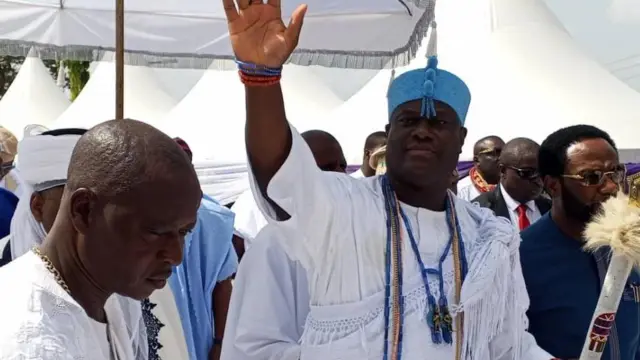 Ooni tí Ife, Ọba Adeyeye Ogunwusi to wọ aṣọ agbada àti Sokoto aláwọ̀ funfun pẹlu ade aláwọ̀ búlúù ń ju ọwọ́ sáwọn èèyàn tó ń kí I, àwọn ẹmẹ̀wà rẹ rọgba yí I ká.