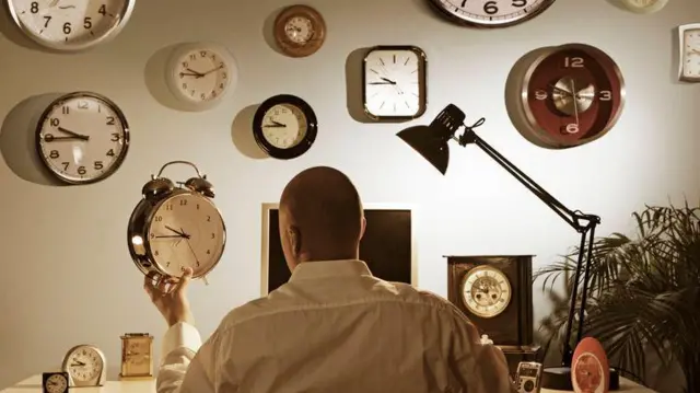 Homem olhando para despertador com vários outros relógios na parede e ao redor dele