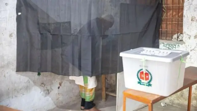 একটি টেবিলের ওপর ব্যালট জমা দেওয়ার বাক্স রাখা। পেছনে কালো রংয়ের স্বচ্ছ পর্দার আড়ালে ভোট দিচ্ছেন টুপি-পাঞ্জাবি ও লুঙ্গি পরা বয়স্ক একজন ব্যক্তি 