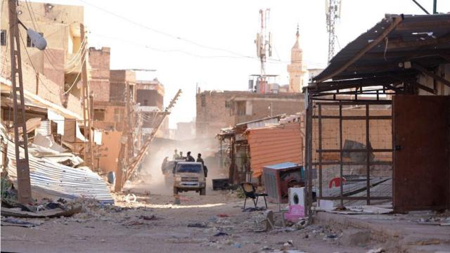 Soldados do exército na traseira de uma caminhonete dirigindo por uma rua destruída em Omdurman