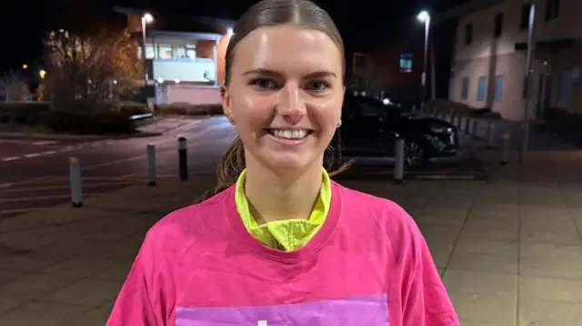 Ella Kempshall lleva una camiseta rosa con el logo de Why, de la organización benéfica contra el cáncer. Tiene el pelo castaño recogido con raya al medio en una coleta baja. Está de pie en una amplia acera junto a unas farolas de noche.