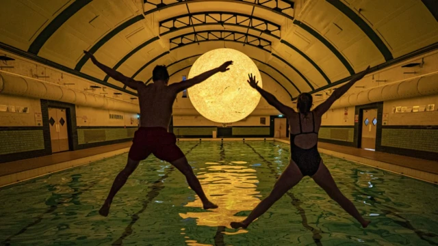 The sun sculpture hanging over a swimming pool with two people, seen from behind, jumping into the water doing star jumps