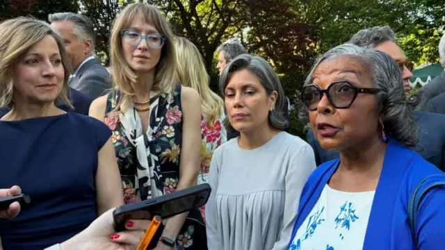 木立を背に、青いジャケットと白いブラウスの黒人女性が取材に答えている。周りに女性が3人立ち、その後ろには大勢の男女がいる。