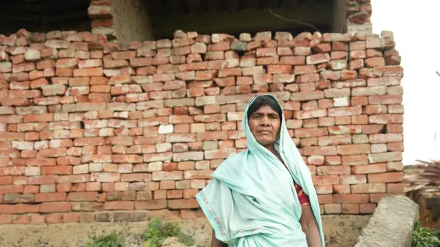 राजो देवी अपने टूटे हुए घर के पास खड़ी हैं