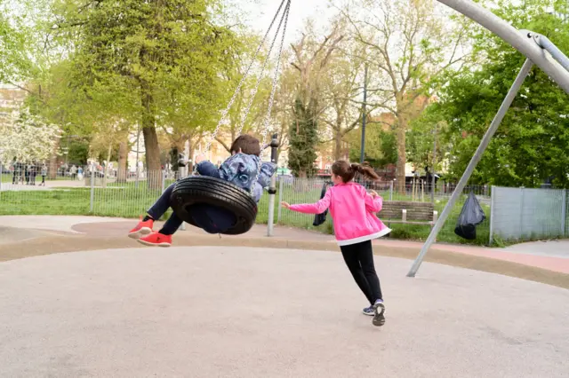 طفلة ترتدي سترة بلون وردي غامق وبنطالاً أسود، شعرها بني وطويل تربطه إلى الخلف، تلعب مع طفل يتأرجح مرتدياً سترة زرقاء مزركشة وبنطالاً يبدو كحلياً وشعره قصير ولونه بني. في حديقة محاطة بالأشجار خلال النهار.