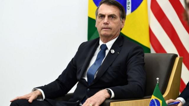 Bolsonaro sentadobonus lampions betuma cadeira, com bandeiras do Brasil e EUA ao fundo