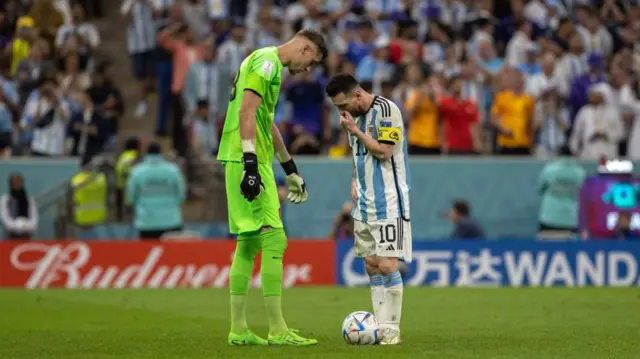 Ліонель Мессі стоїть на футбольному полі у синьо-білій футболці навпроти воротаря збірної Нідерландів Андріса Нопперта. Голландський гравець у зеленій футболці височіє над своїм суперником. На задньому плані розмита аудиторія.