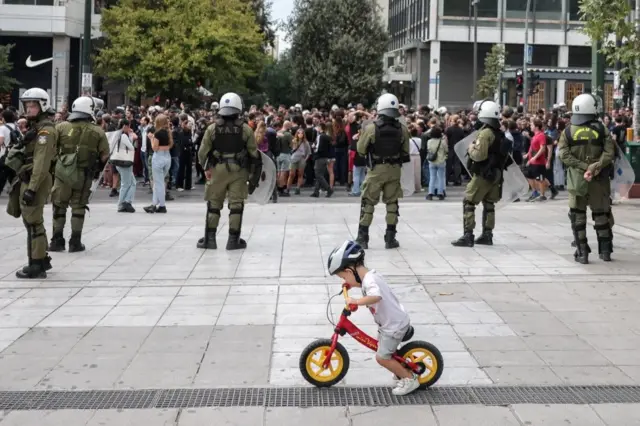 Dete vozi bicikl dok policija za suzbijanje demonstracija stoji na straži tokom demonstracija povodom 24-časovnog generalnog štrajka u Atini protiv reforme kojom se produžava 13-časovno radno vreme za radnike sa jednim poslom