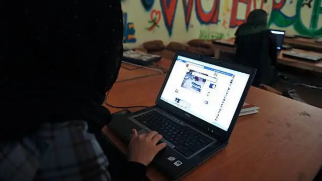 تصویر زنی که در مقابل کمپویترش نوشته و شبکه‌های اجتماعی را در کمپوتر مرور می‌کند. چهره زن معلوم نیست پشت او به سمت دوربین است. در صفحه کمپیوتر شبکه اجتماعی فیسبوک دیده می‌شود. 