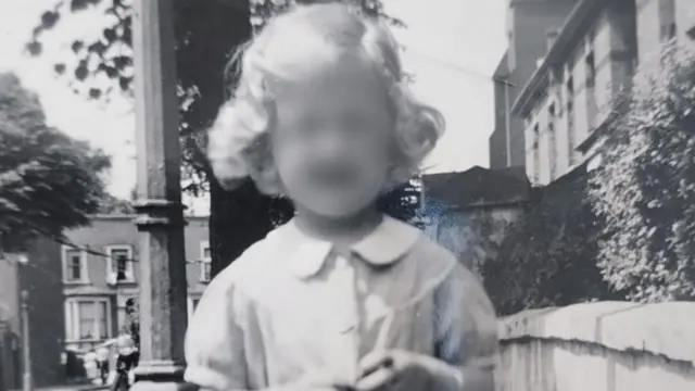Fotografia em preto e branco de Susan (não é seu nome verdadeiro) tirada nos anos 1950 ou 1960. Ela aparenta ter cerca de sete anos de idade e está em pé em uma rua com casas geminadas atrás dela e um muro à sua esquerda. Ela tem longos cabelos loiros e está usando um vestido.