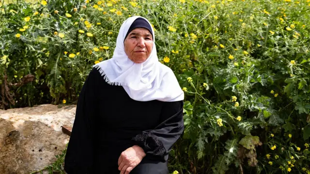 Ayesha Shtayyeh,jogar grátis caça niquel bartúnica preta e um lenço branco na cabeça, com uma expressão séria no rosto, sentadajogar grátis caça niquel barfrente a uma plantaçãojogar grátis caça niquel barflores amarelas.