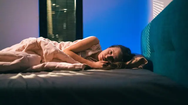 Stresszt és nyugtalanságot tükröző, nyitott szemmel fekvő nő egy sötét hálószobában.