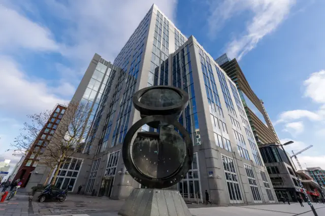 欧洲清算银行的高耸现代化办公大楼,外墙为反光玻璃窗,照片从低角度拍摄,前景有一个大型圆形金属雕塑,天空湛蓝并散布白云