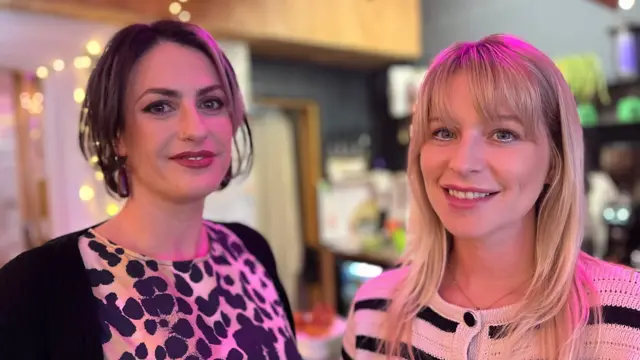 Madelaine Thomas e Jess Davies em uma cafeteria, sorrindo para a câmera. Thomas tem cabelo curto e veste uma camiseta com estampa de animal e um casaco preto. Davies tem cabelo loiro comprido com franja e veste um casaco listrado preto e branco.