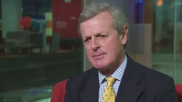 Седой мужчина в темном пиджаке, рубашке в голубую клетку и желтом галстуке внимательно слушает вопрос интервьюера