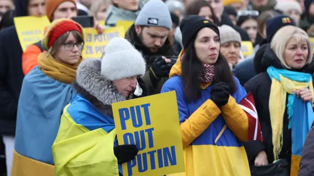 Жители Риги митингуют в годовщину начала полномасштабного вторжения России в Украину. Они стоят, завернувшись в украинские флаги 