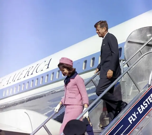 Jackie Kennedy descendo as escadas de um avião. Ela veste um conjunto de saia e blazer rosa, com chapéu rosa. Atrás dela está o marido, Jhon Kennedy, de terno.
