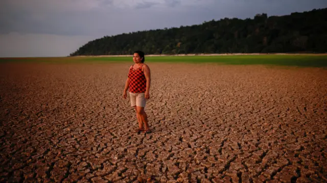 ஒரு பெண் வறண்ட மற்றும் வெடித்த ஆற்றுப் படுக்கையில் நிற்கிறார், தூரத்தில் ஒரு மெல்லிய நீரோட்டம் தெரிகிறது.