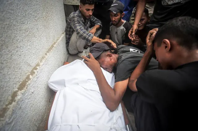 أقارب لفلسطينيين لقوا حتفهم، نتيجة للهجمات الإسرائيلية على أجزاء مختلفة من مدينة غزة، ينعون أقاربهم الموتى في مستشفى الشفاء خلال إجراءات جنازتهم في 27 أغسطس/آب 2025، وتظهر في الصورة جثة مغطاة بالكفن الأبيض، بينما يبكي شخص بجوارها محاطاً بأشخاص أخرين يواسوه.