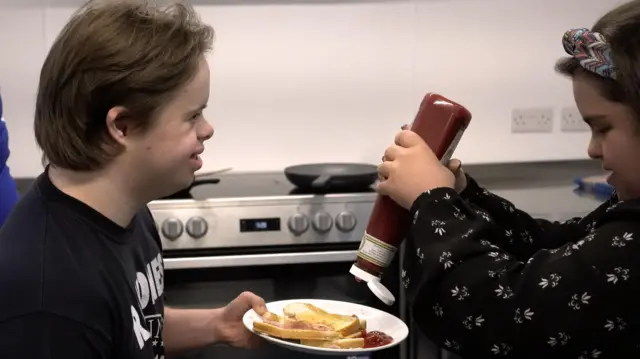 Dois jovens com síndromeprognóstico de futebol jogos de hojeDown, um deles segurando um prato com sanduíche, e o outro colocando ketchup no prato