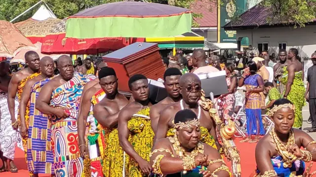 The king's gold treasures, carried in a box, are paraded before the crowd by a group of men walking and women dancing