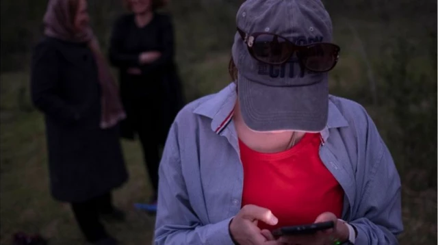 Une personne tête baissée, regarde son téléphone. Son visage est caché par la casquette qu'elle porte.